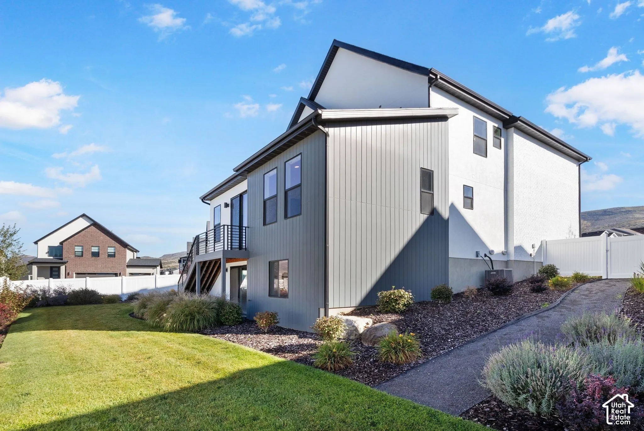 Modern exterior featuring vertical siding, a white stucco finish, a second-story deck with metal railings, a manicured lawn, and a paved pathway