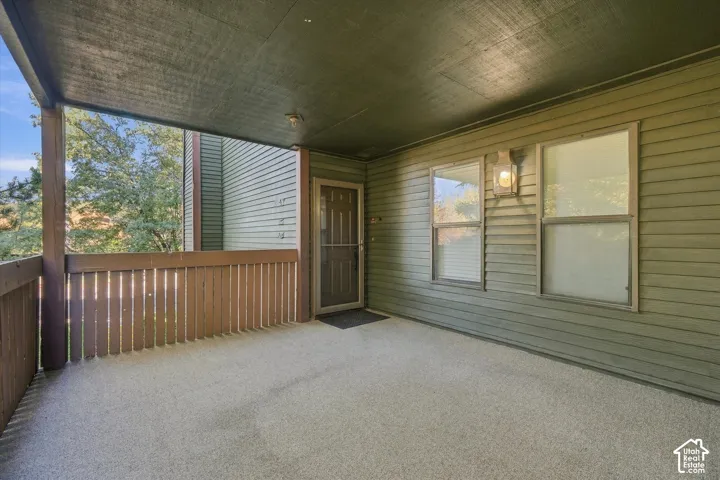 Covered outdoor patio with a textured flooring surface, wooden railing, and a wall-mounted light fixture