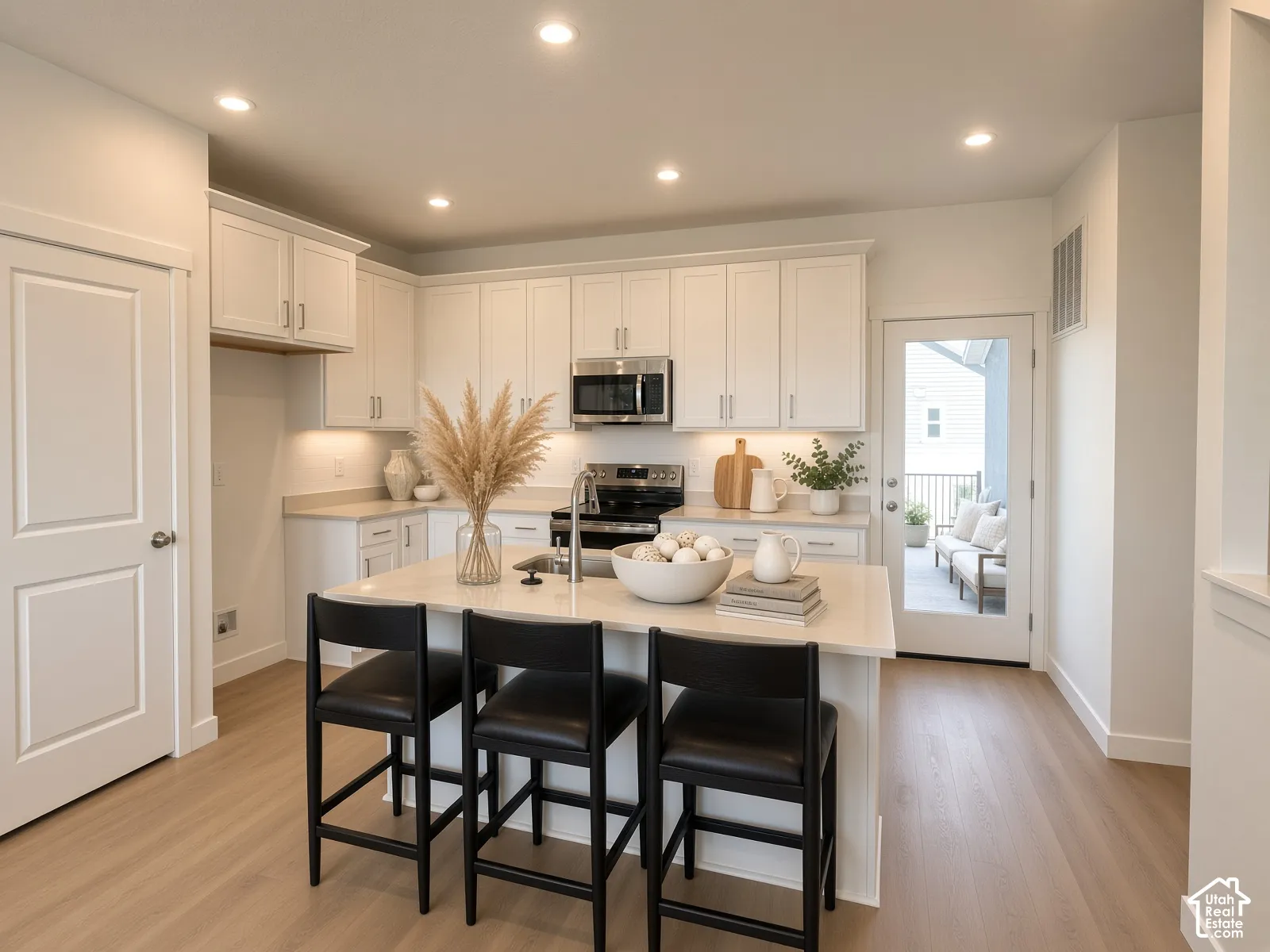 Light-toned wood-finish flooring, white cabinetry, recessed lighting, stainless steel appliances, and a glass-paneled door leading to an outdoor deck