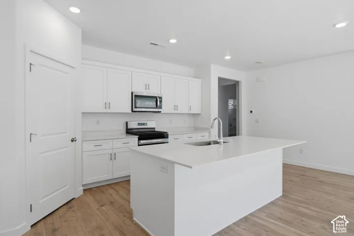 Kitchen featuring white cabinetry, stainless steel appliances, a center island with an integrated sink, and wood-finish flooring