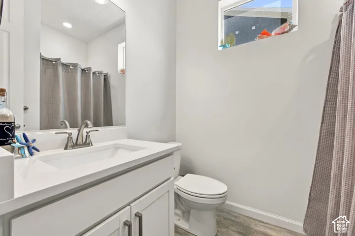 Vanity with a single basin sink and brushed nickel faucet, large wall mirror, toilet, wood-finish flooring, and a high-set window