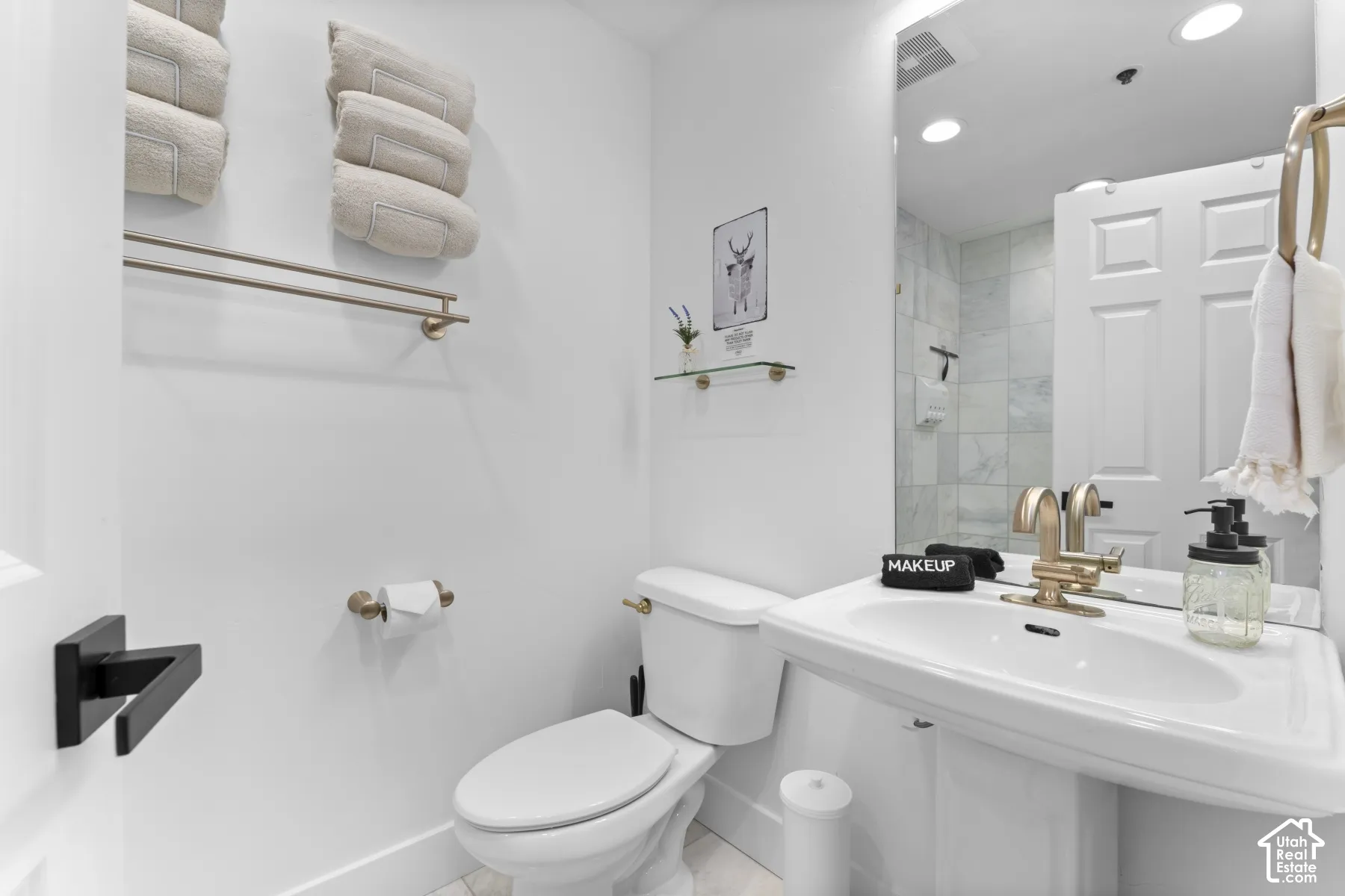 Pedestal sink with a gold-tone faucet, wall-mounted mirror, white toilet, and marble-patterned shower tile
