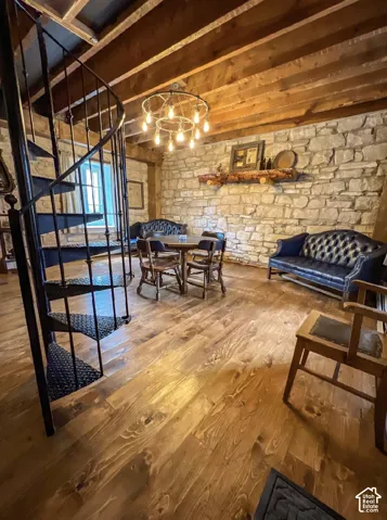 Stone accent wall, exposed wood beam ceiling, wood-finish flooring, black spiral staircase, and a wagon wheel chandelier
