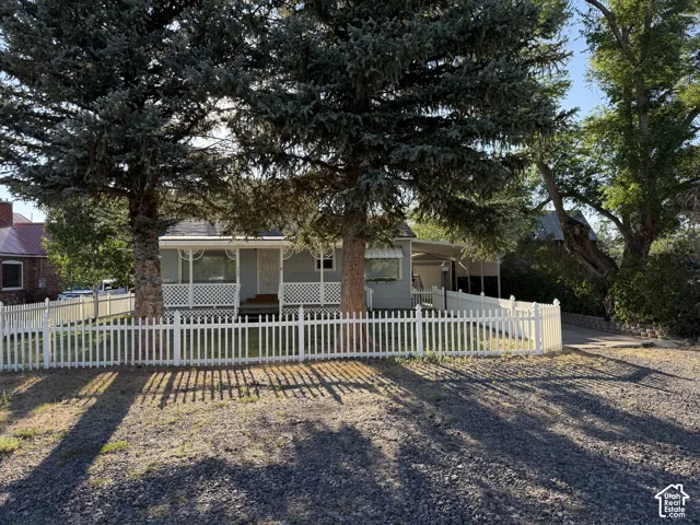 Front exterior featuring a white picket fence, mature evergreen trees, a covered porch, and a gravel driveway