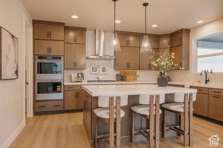 Kitchen featuring wood-finish cabinetry, a large central island with a solid surface countertop, stainless steel double wall ovens, a stainless steel range hood, and glass pendant lighting