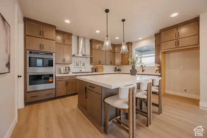 Kitchen featuring a central island with quartz countertops, wood-finish cabinetry, stainless steel double wall ovens, a chimney-style range hood, and pendant lighting