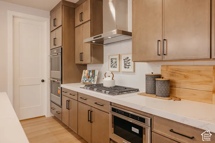 Kitchen featuring stained wood cabinetry, stone-finish countertops, stainless steel wall-mounted range hood, built-in double wall ovens, and gas cooktop