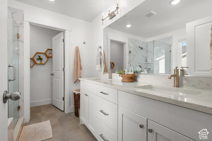 Dual vanity with integrated sinks and brushed brass fixtures, white cabinetry with silver-tone hardware, large vanity mirror, glass-enclosed shower with tiled walls, and recessed lighting