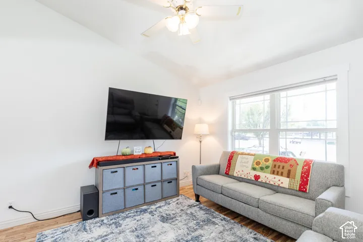 Vaulted ceiling with a ceiling fan light fixture, wood-finish flooring, and a large window with white trim