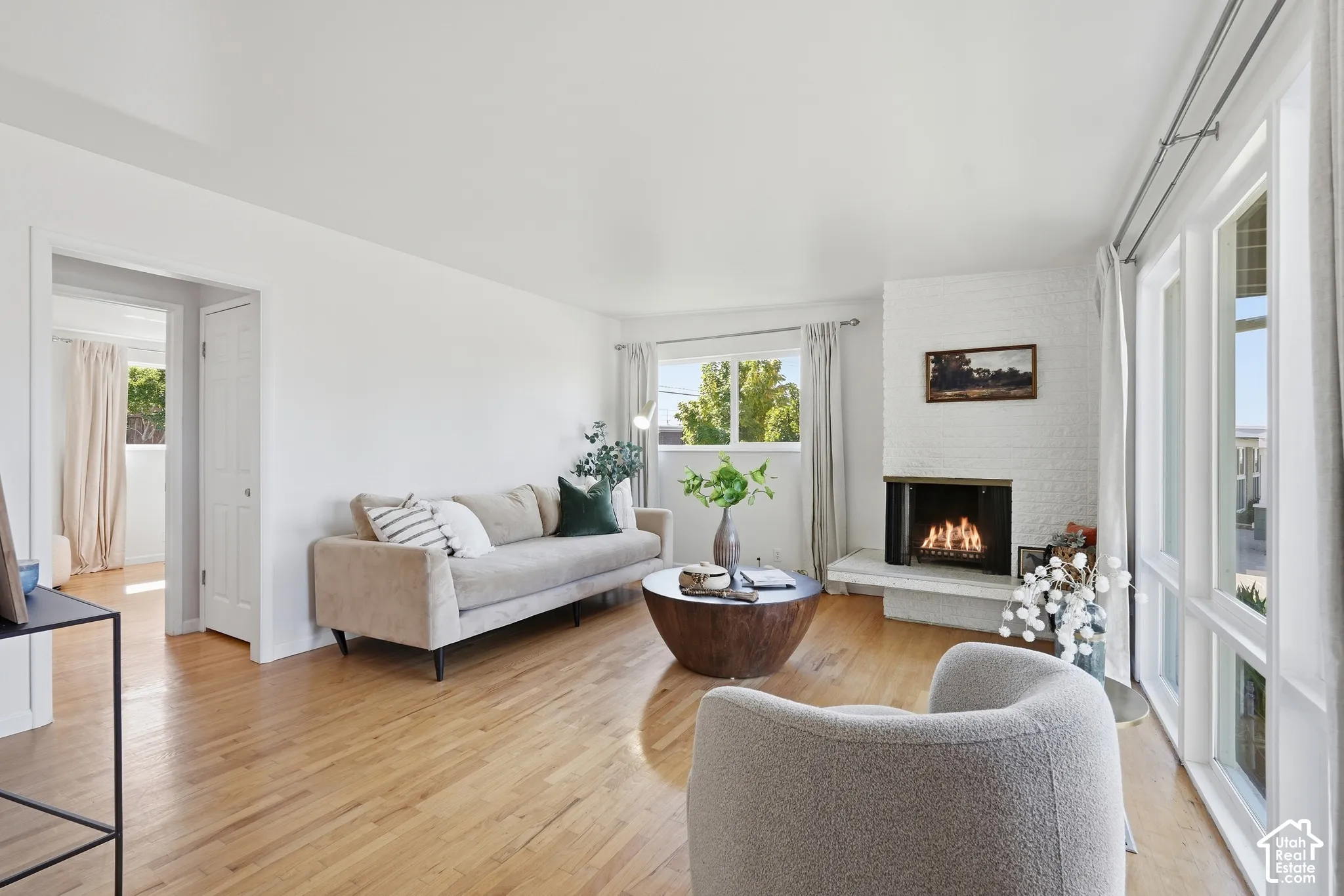 Light wood-finish flooring, white painted walls, white brick fireplace, multiple windows with white trim, and a white interior door