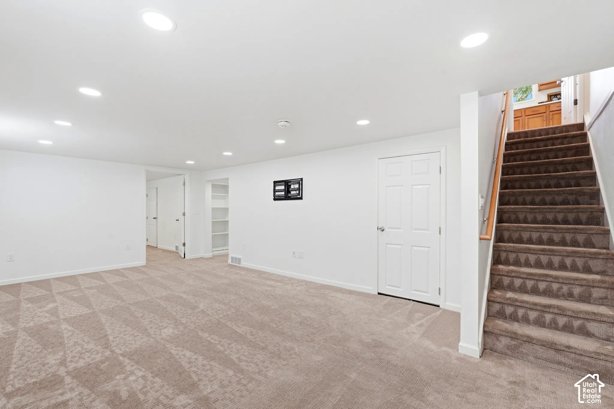 Recessed lighting illuminates the carpeted room, featuring a built-in storage unit, a paneled door, and a staircase with a wood handrail