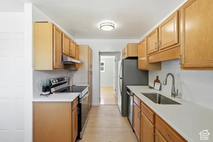 Galley kitchen featuring wood-finish cabinetry, stainless steel appliances, light-toned countertops, subway tile backsplash, and a ceiling-mounted light fixture