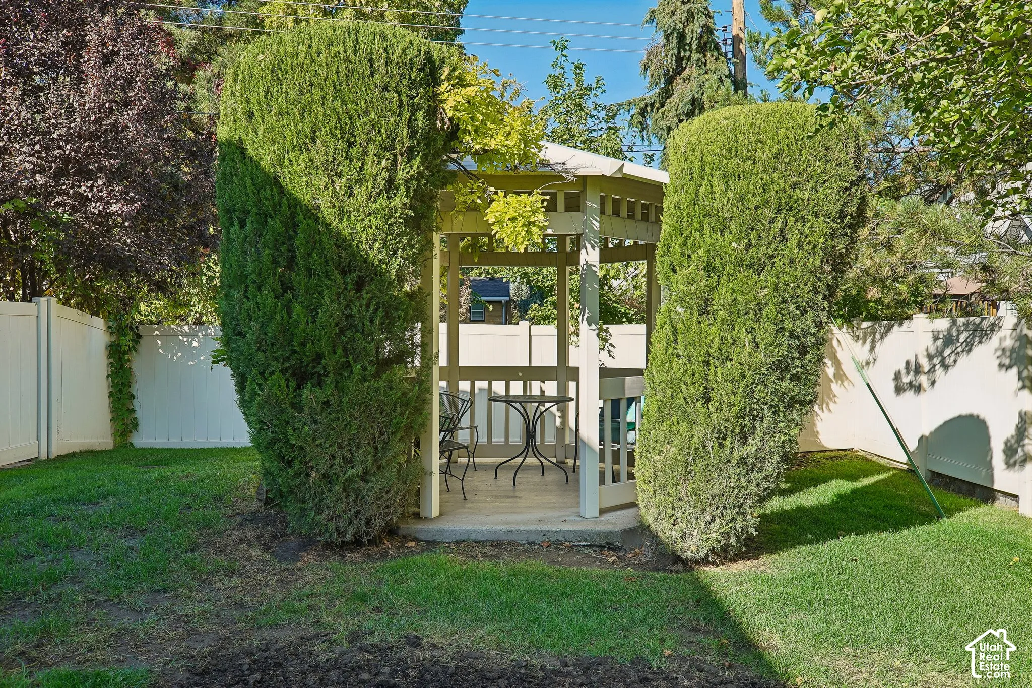 Outdoor gazebo structure with a built-in bench, surrounded by manicured hedges and a white privacy fence