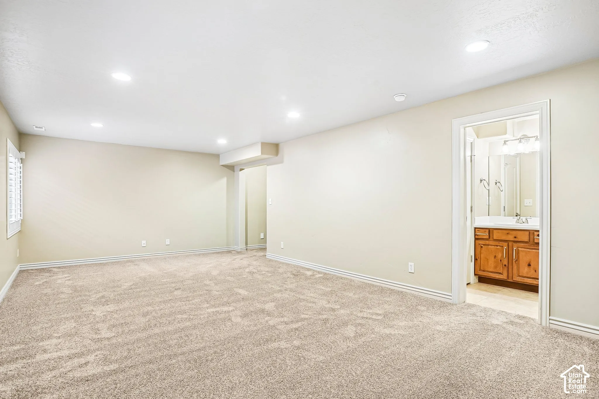 Recessed lighting, wall-to-wall carpeting, window with plantation shutters, and an adjacent bathroom featuring a wood-finish vanity with a built-in sink