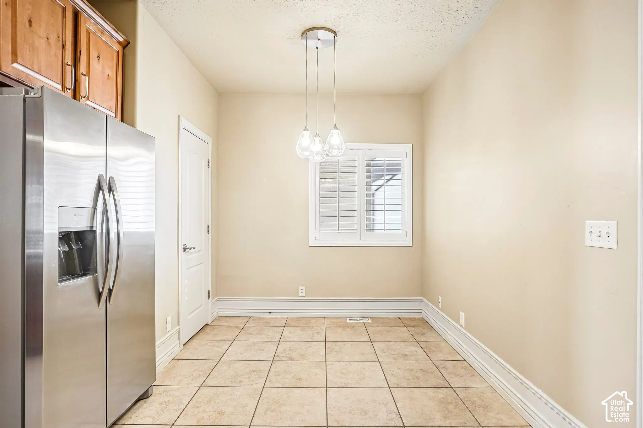Spacious kitchen featuring stainless steel side-by-side refrigerator with ice and water dispensers, neutral tile flooring, soft ambient pendant lighting, and abundant natural light from shuttered window