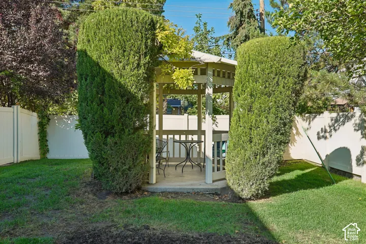 Outdoor gazebo structure with a built-in bench, surrounded by manicured hedges and a white privacy fence