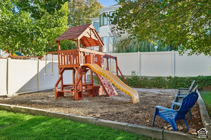 Outdoor play area featuring a multi-level playset with slide and swings, wood chip ground cover, and a surrounding white privacy fence
