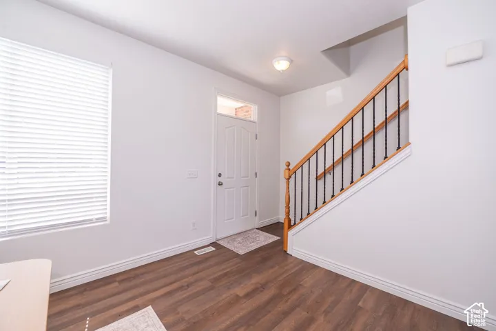 Entryway featuring wood-finish flooring, a staircase with wooden handrails and wrought iron balusters, a white paneled entry door with sidelight, and a large window with blinds