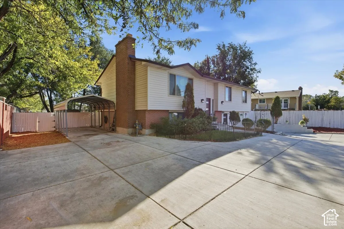 Extended concrete driveway with a metal carport structure, brick chimney, and mature trees