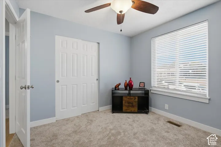 Plush carpeted flooring, a large window with horizontal blinds, a ceiling fan with integrated lighting, and a sliding door closet