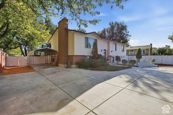 Extended concrete driveway with a metal carport structure, brick chimney, and mature trees