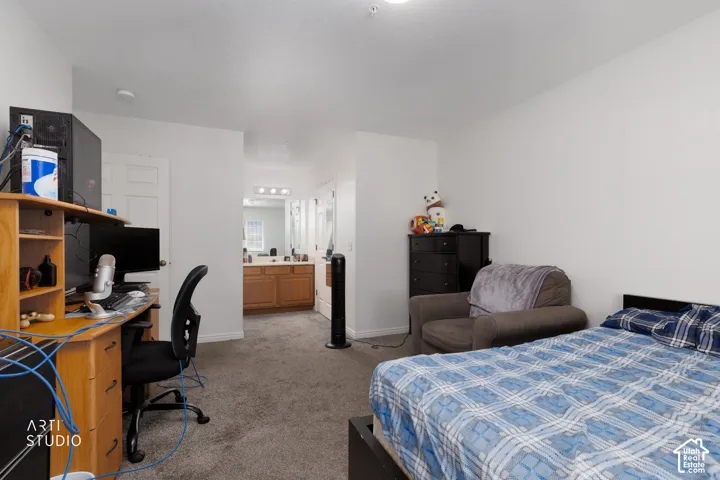 Carpeted room with white walls, featuring a built-in desk with shelving and a dedicated bathroom area with a wood-finish vanity and multiple mirrors