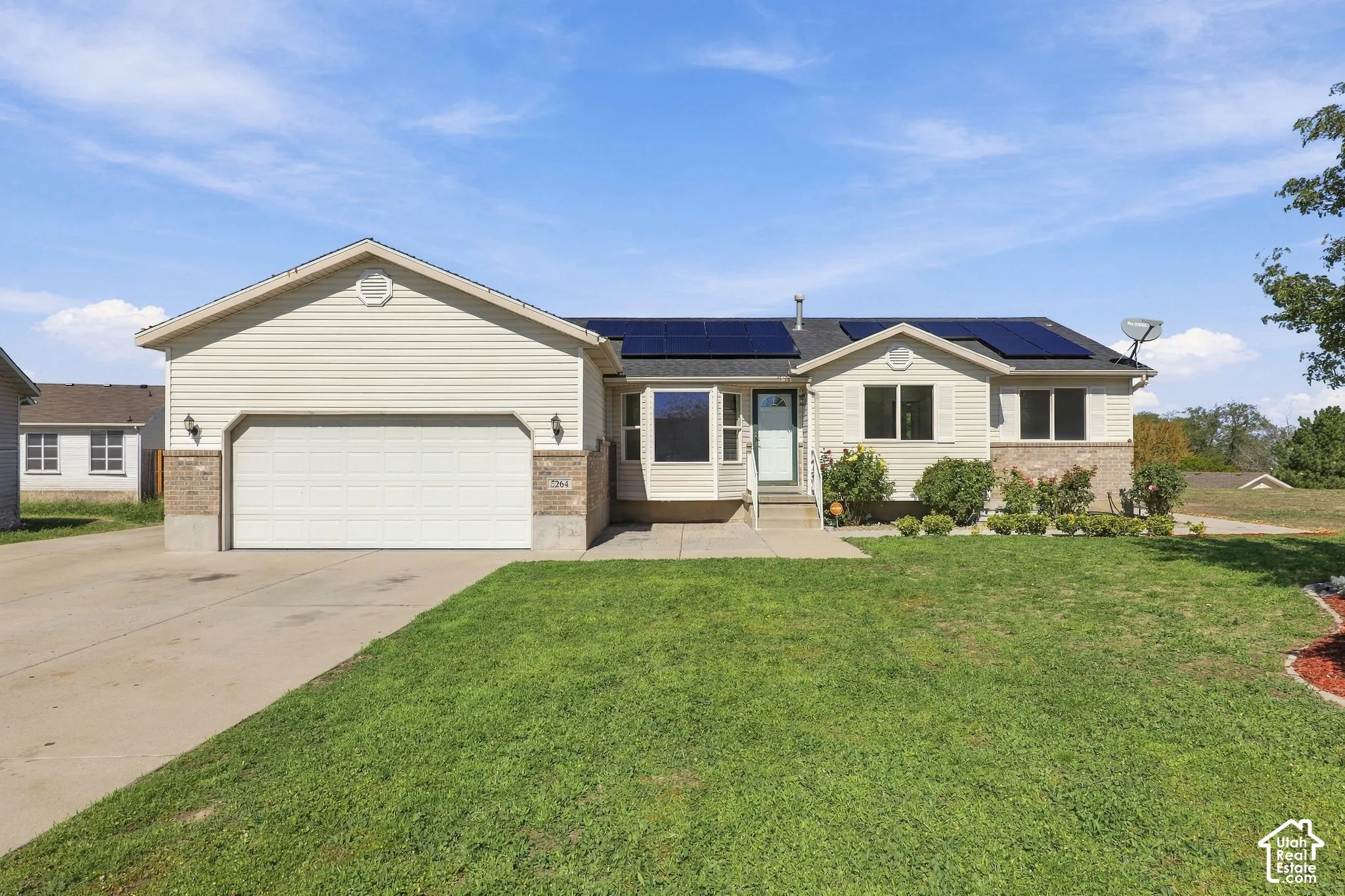 Exterior featuring light-colored siding, partial stone-finish facade, an attached garage with a drive, solar panels on the roof, and established landscaping