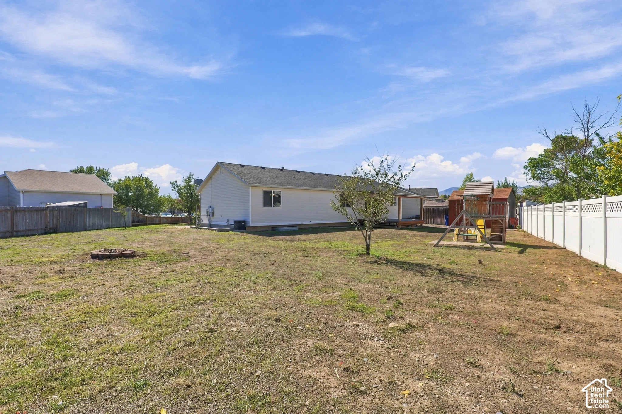 Expansive backyard featuring a white vinyl privacy fence, a Playground, a small tree, and a built-in fire pit