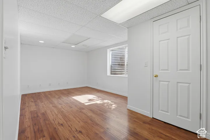 Wood-finish flooring, drop ceiling with recessed lighting, white panel door with brass hardware, and a window with a security well