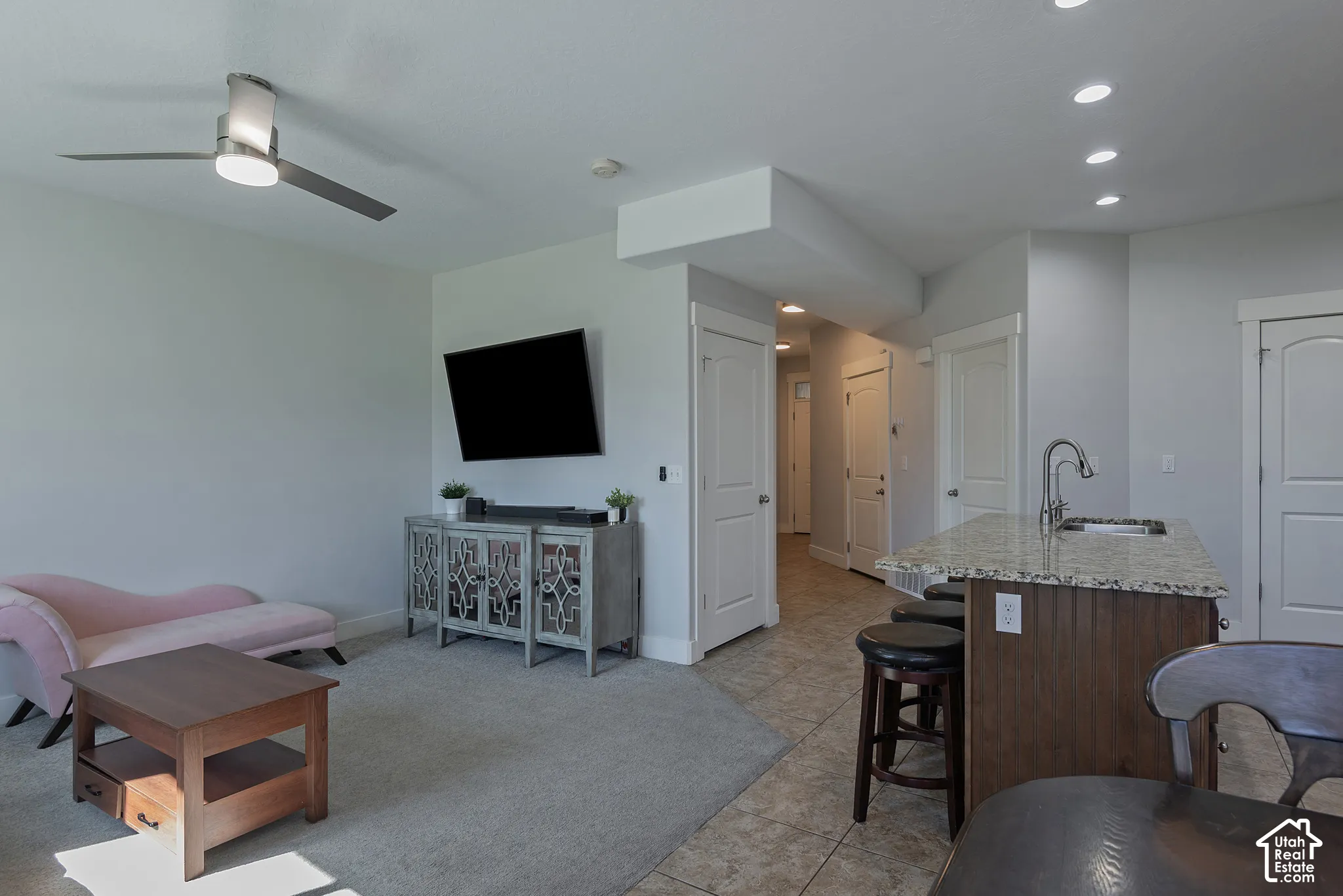 Open-concept living space featuring a ceiling fan with integrated lighting, wall-mounted television, and a kitchen island with stone-finish countertop and sink