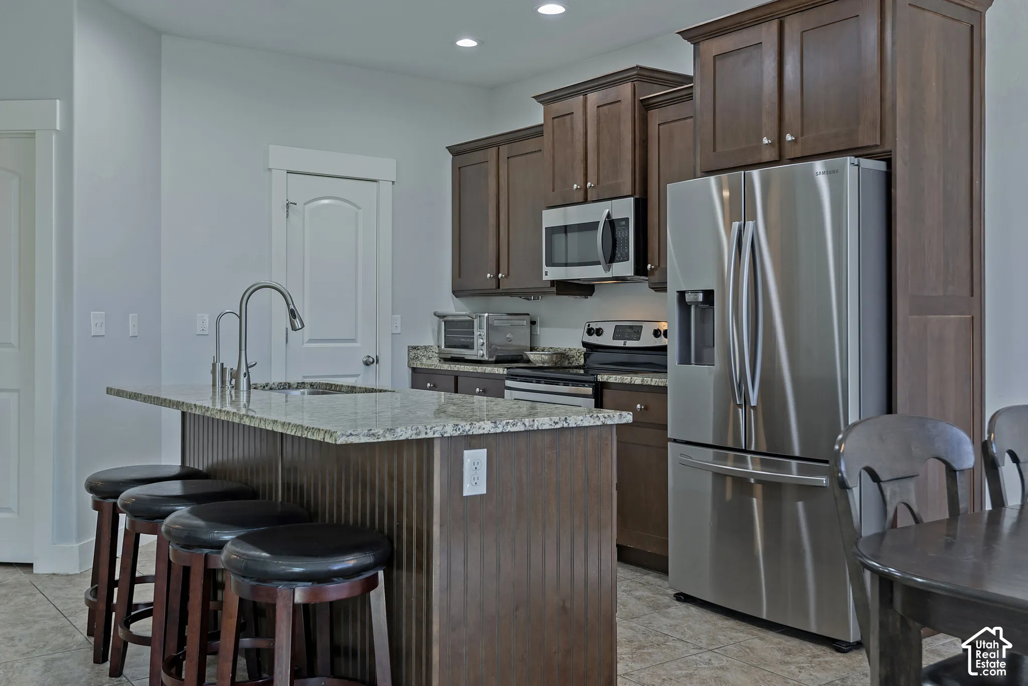 Kitchen featuring wood cabinetry, stainless steel appliances, a granite-top island with a sink, and tile flooring