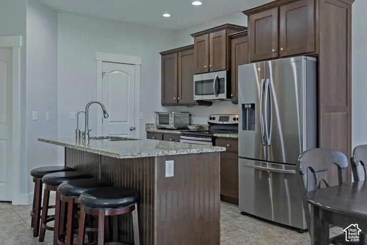 Kitchen featuring wood cabinetry, stainless steel appliances, a granite-top island with a sink, and tile flooring