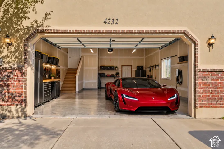 Multi-car garage featuring epoxy-coated flooring, built-in metal cabinetry with workspace, recessed ceiling lighting, wall-mounted electric vehicle charging station, and staircase with wood treads
