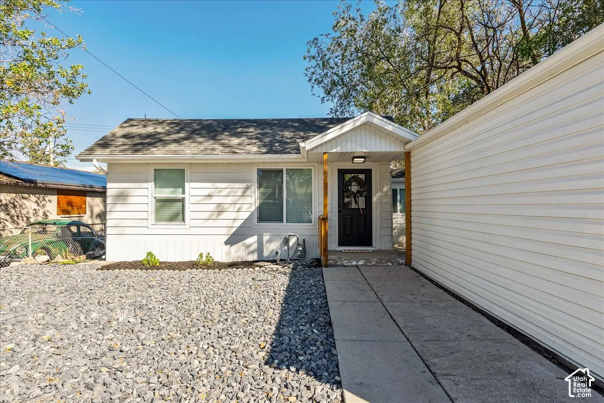 White siding exterior, dark entry door with overhead lighting, concrete walkway, gravel landscaping, and a side-mounted accessory structure