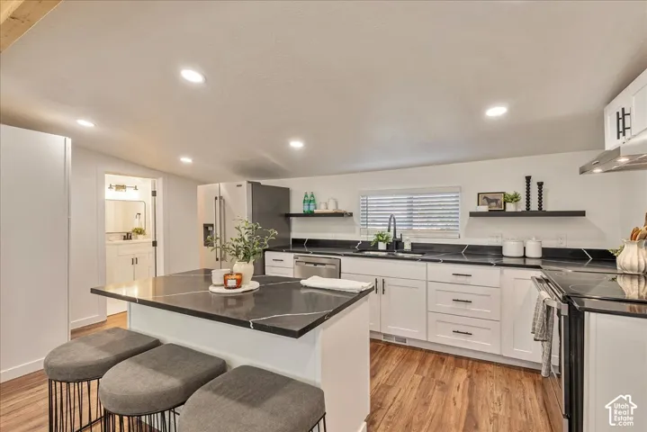 White cabinetry with dark hardware, wood-finish flooring, recessed lighting, kitchen island with seating, and stainless steel appliances