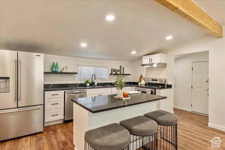 Modern kitchen featuring stainless steel appliances, dark countertops, white cabinetry, wood-finish flooring, and a central island with bar seating