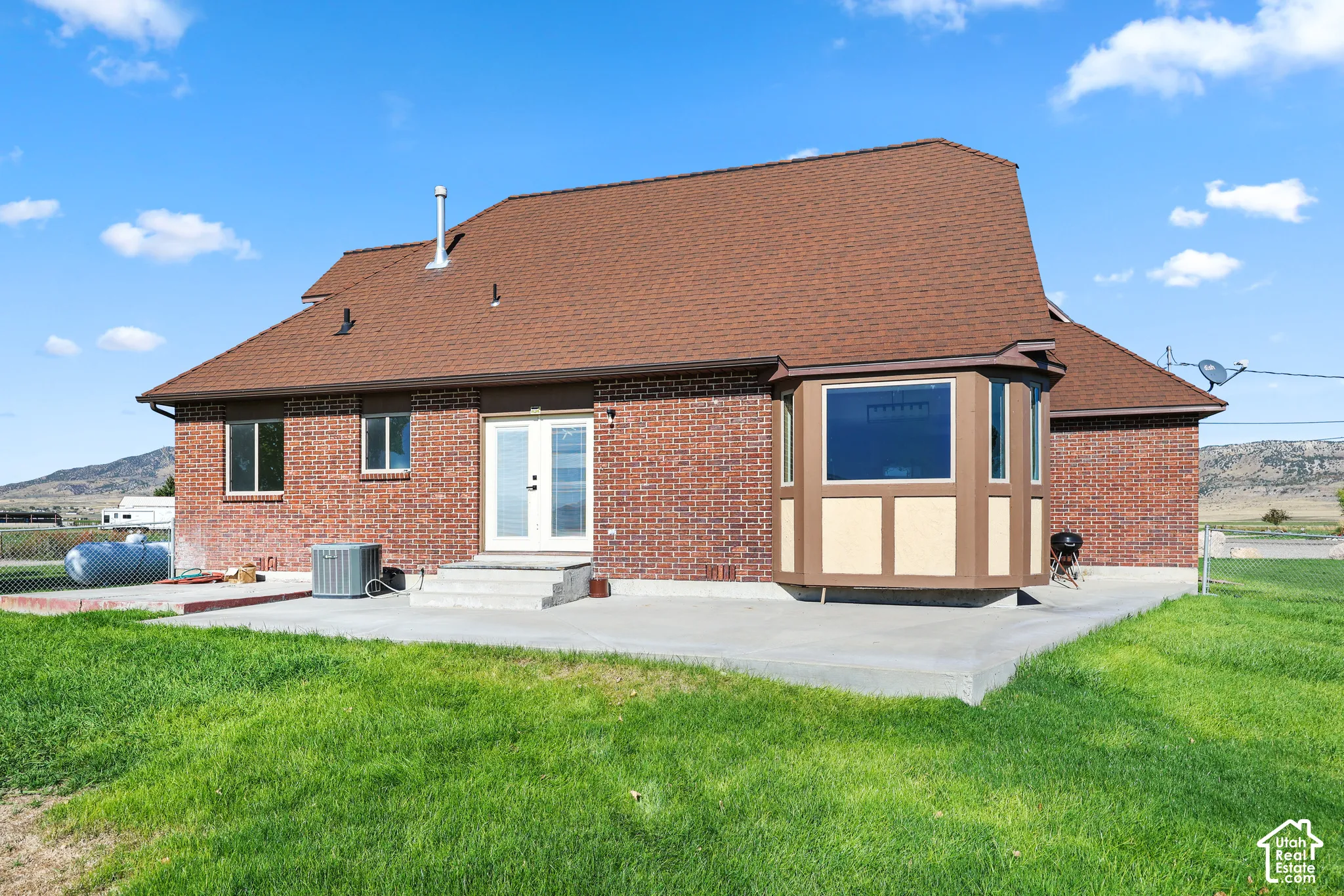 Brick exterior with a bay window, concrete patio, double glass-paneled doors, and a brown shingle roof