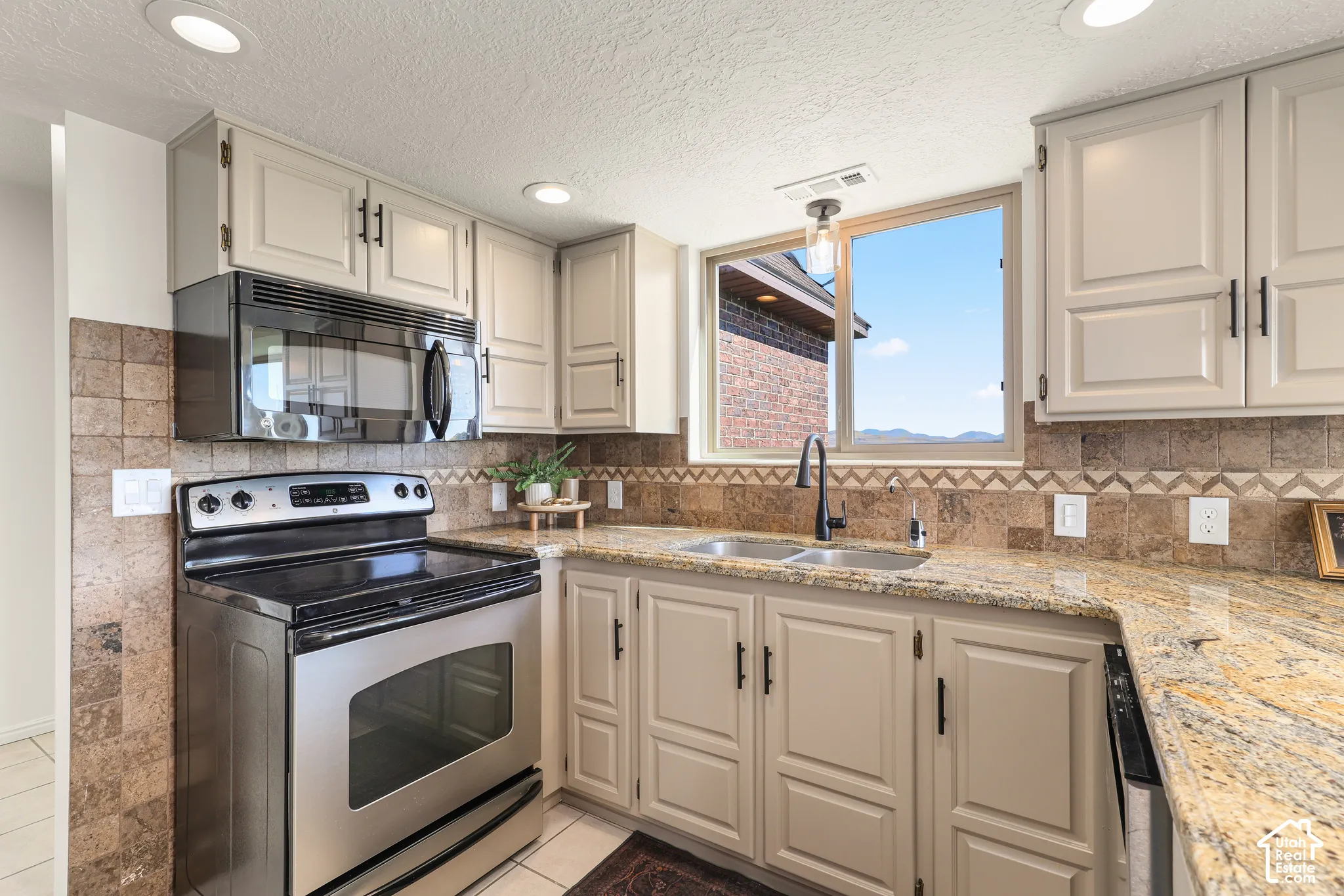 Light-toned cabinetry with black hardware, stainless steel appliances, stone-finish countertop, tile backsplash with decorative accent, and a stainless steel double basin sink with a black faucet