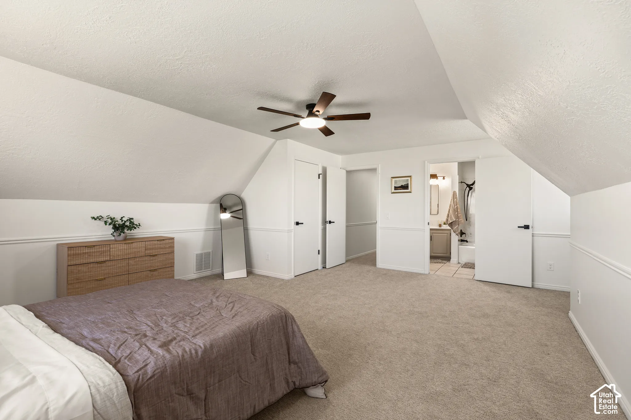 Angled ceiling lines with a ceiling fan, carpet flooring, white painted walls, and an attached bathroom featuring a vanity with a rectangular sink