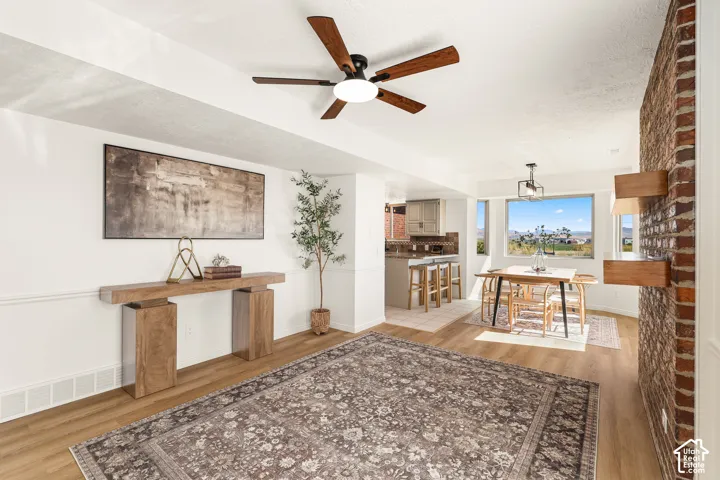 Wood-finish flooring throughout, a modern ceiling fan, an exposed brick accent wall with floating wood shelves, wainscoting, and large windows