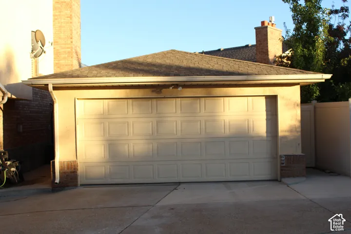 Attached garage with a panel-style door, finished stucco exterior, brick base detailing, and a shingle roof