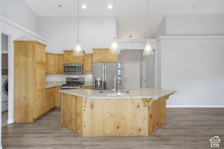 Kitchen featuring wood cabinetry, stainless steel appliances, a center island with a sink, pendant lighting, and wood-finish flooring
