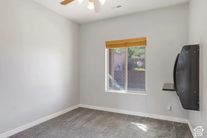 Neutral wall paint, gray carpet flooring, window with wood-finish blinds, ceiling fan with integrated lighting, and a wall-mounted shelf unit