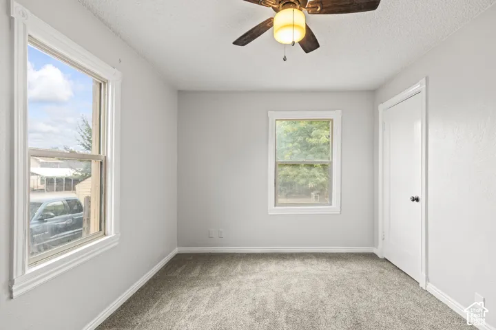Neutral gray wall paint, textured carpet flooring, two windows with white trim, and a ceiling fan with integrated lighting