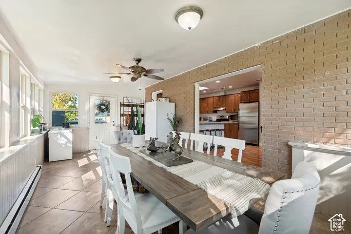 Open concept living area with brick-finish accent wall, ceiling fan, multiple windows, and tiled flooring