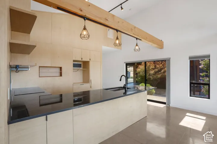Sleek kitchen island with a dark countertop and undermount sink, light wood-finish cabinetry, exposed wood beam with track lighting, and a vaulted ceiling