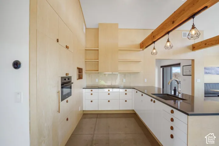 Kitchen featuring light wood-finish cabinetry, white lower cabinets with circular wood pulls, dark stone countertops, a stainless steel undermount sink with a gooseneck faucet, and exposed wood beams with pendant lighting