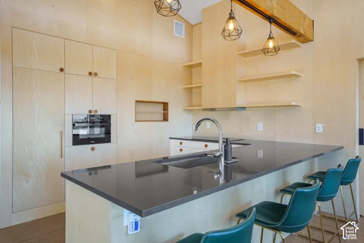 Kitchen featuring light wood-finish cabinetry, built-in oven, island with dark countertop, integrated sink, and exposed beam accent