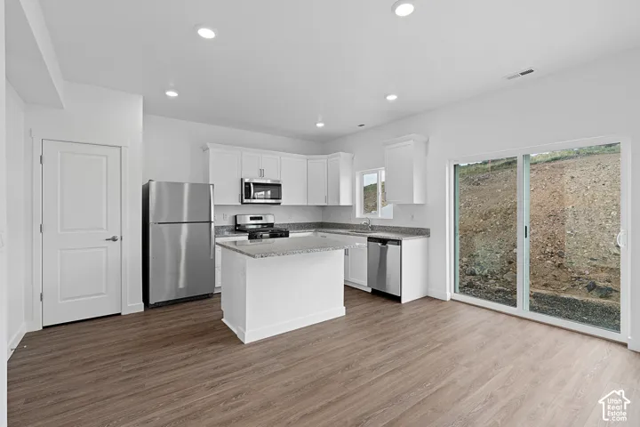 Open-concept kitchen featuring white cabinetry, stainless steel appliances, a center island with a granite-style countertop, and wood-finish flooring