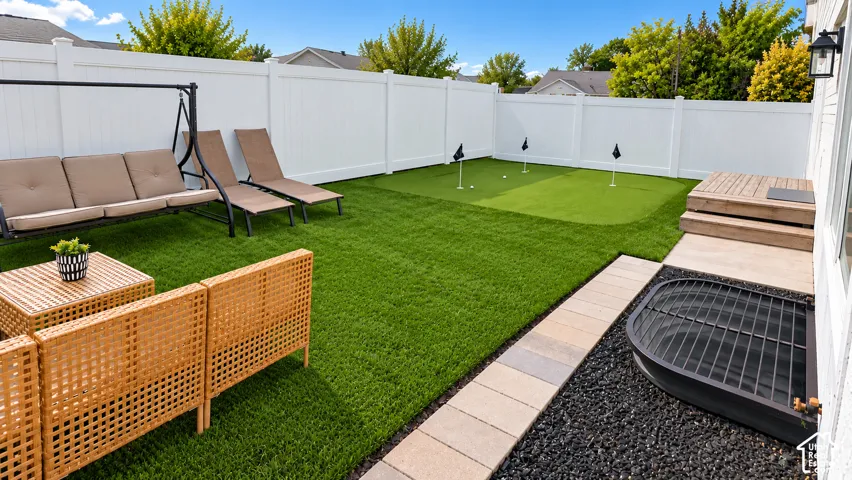 Expansive artificial turf backyard featuring a putting green, white privacy fencing, a raised wood deck, and a paver stone walkway